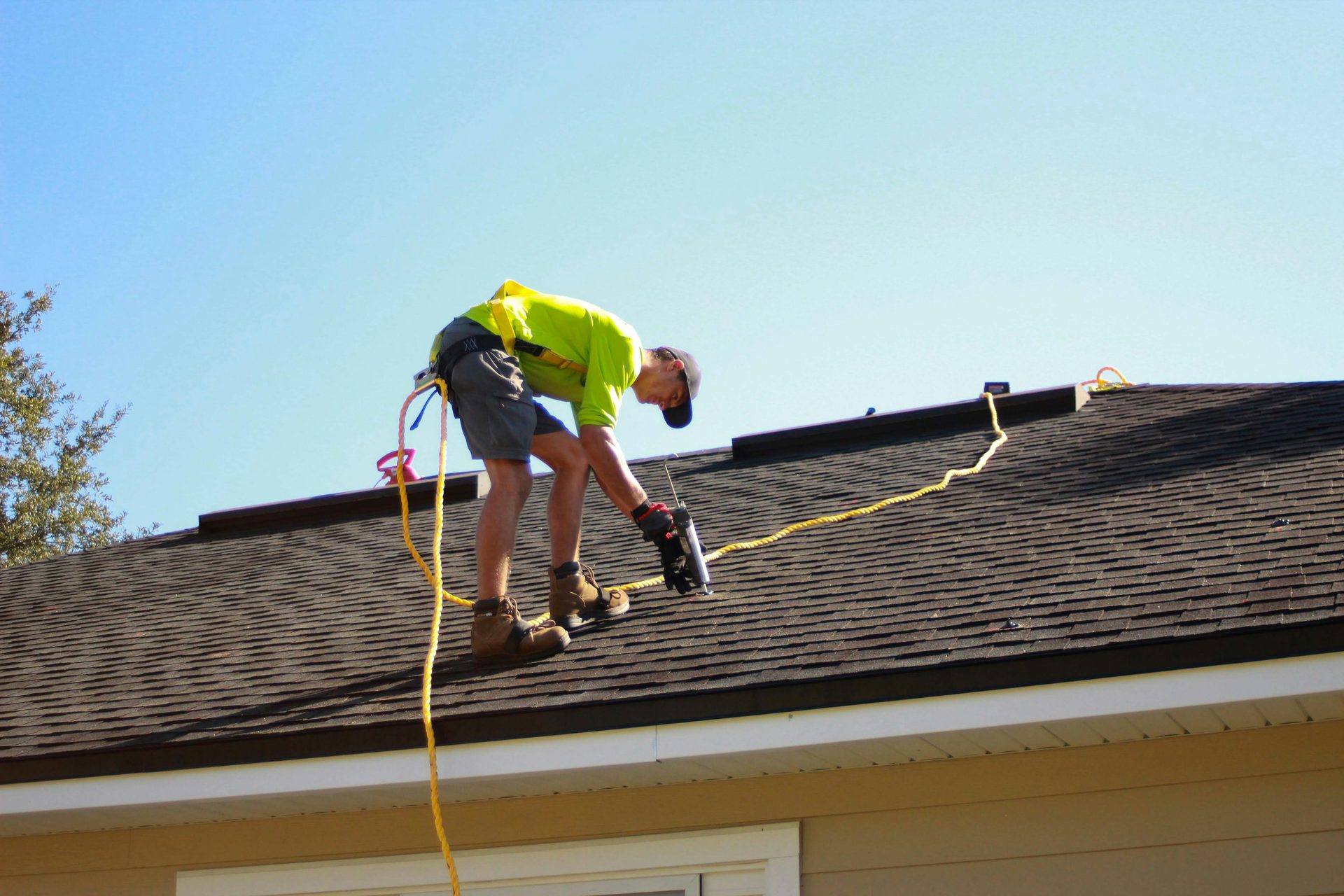 A roofer in a harness fastening shingles on a pitched roof.